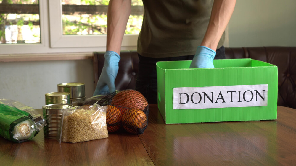Volunteer in the Protective Medical Mask and Hand Gloves Putting Food in Donation Box.